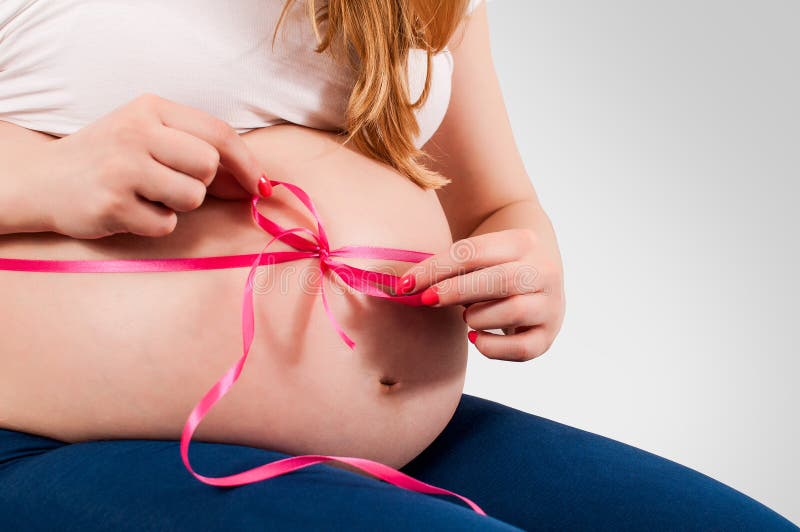 Pregnant girl tying pink bow at her belly. Pregnant girl sitting on the sofa and tying pink bow at her belly, horizontal image royalty free stock photo