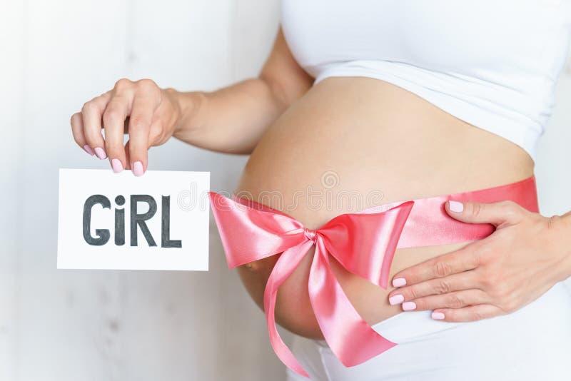 Tummy with pink ribbon and bow of pregnant caucasian woman which is holding nameplate girl. In light room stock photos
