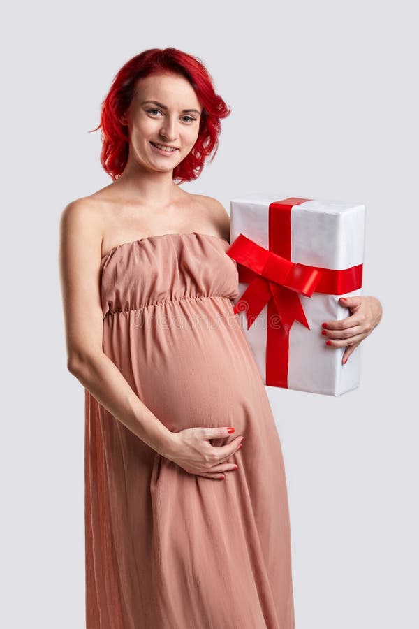 A pregnant girl is holding a large white gift box with a red ribbon and bow. Studio. A young red-haired pregnant girl in a long light dress is standing and is royalty free stock photo